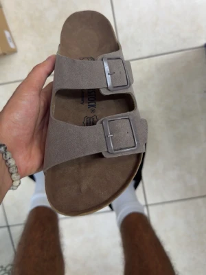 Birkenstock sandaler - Säljer ett par klassiska Birkenstock Arizona sandaler i beige mocka med två breda remmar och justerbara metallspännen. Fotbädden är ergonomiskt utformad och sulan är i kork och gummi för extra komfort. Perfekt för dig som gillar stilren och avslappnad look.