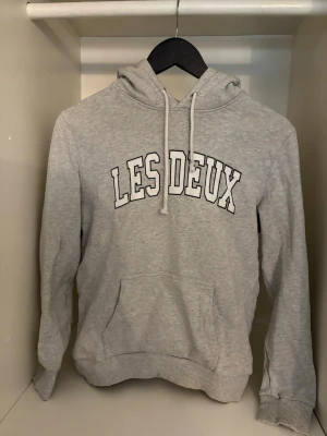 Grå hoodie från Les Deux - Snygg grå hoodie från Les Deux med stor vit logga på bröstet. Klassisk modell med huva och dragsnören samt en stor magficka. Tillverkad i mjukt sweatshirtmaterial och har ribbade muddar vid ärmslut och nederkant. Perfekt för en avslappnad streetwear-look.