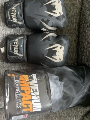 Venum Impact boxningshandskar svart - Säljer ett par svarta Venum Impact boxningshandskar med coolt kamouflagemönster och vita loggor. Handskarna har kardborreband för enkel justering och är tillverkade i slitstarkt syntetmaterial. Perfekta för träning och sparring.
