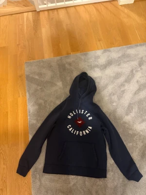 Mörkblå hoodie från Hollister - Mörkblå hoodie från Hollister med vit text 'Hollister California' och röd logga på bröstet. Klassisk känguruficka framtill och justerbar huva. Perfekt för en avslappnad och stilren look. Står XL men passar L
