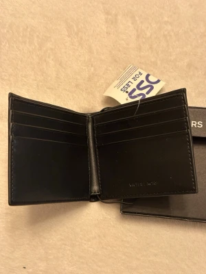 Svart plånbok från Michael Kors - Stilren svart plånbok från Michael Kors i läder med präglad struktur. Plånboken har flera kortfack och ett större fack för sedlar. Diskret logga i silver på framsidan och insidan. Perfekt för dig som vill ha en klassisk och snygg accessoar. Oanvänd i förpackning. Nypris 1000kr