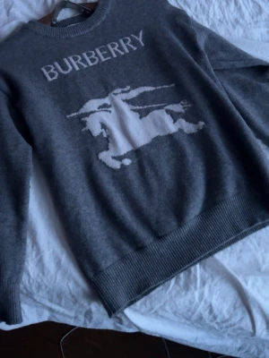 Grå stickad tröja från Burberry - Snygg grå stickad tröja från Burberry med vit logotyp och klassiskt riddarmotiv framtill. Tröjan har rund halsringning och ribbade muddar vid ärmslut och nederkant. Perfekt för dig som gillar stilrena och exklusiva plagg.