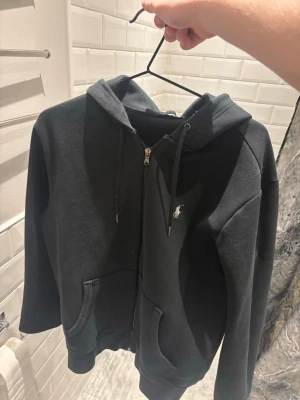 Svart zip hoodie från Ralph Lauren - Snygg svart hoodie med dragkedja från Ralph Lauren. Klassisk design med huva och dragsnören, två fickor framtill och den ikoniska vita logon broderad på bröstet. Perfekt till chill vardagsstil. Mjuk bomullsblandning för skön känsla.