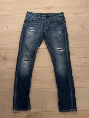 Blå gråa jeans med slitningar  - Säljer ett par blå jack n Jones jeans med slitningar. Snygga Jeans som sitter skönt och ser lika bra ut. Finns ett hål vid gylfen, går lätt att sy igen. Bara att höra av sig vid funderingar