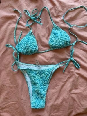 Turkos bikini med smock och knyt - Säljer en turkos bikini med smockdetaljer och knyt i sidorna samt runt nacken och ryggen, från Shein . Bikinin har trekantsformad överdel och klassiska bikinitrosor. Perfekt för stranden eller poolen!  Aldrig använd!