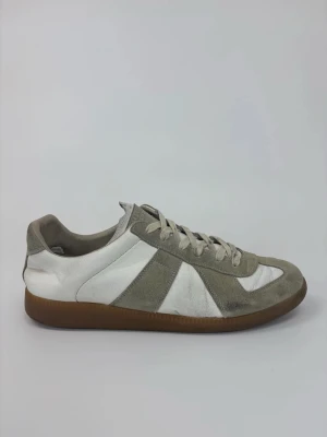 Vita och grå Maison Margiela sneakers med mockadetaljer - Snygga sneakers i vitt och grått med ovandel i läder och mocka. Skorna har klassisk låg profil, vita snören och en brun gummisula som ger retrovibbar. Perfekta för dig som gillar stilrena och tidlösa sneakers.
