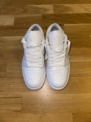 Vita Air Jordan 1 Low sneakers - Säljer ett par helvita Air Jordan 1 Low sneakers från Nike. Skorna har klassisk låg siluett, perforerad tåbox och ikoniska swoosh-loggan på sidan. Tillverkade i läder med vita skosnören och diskret Jumpman-logga på plösen. Perfekta för en clean och stilren look.
