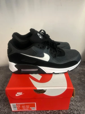Nike Air Max 90 svart/vit sneakers - Nike Air Max 90 sneakers i svart och grått med vit swoosh. Klassisk siluett med mesh och läderdetaljer, synlig Air Max-dämpning i sulan och svarta skosnören. Perfekt för dig som gillar streetwear och sportig stil.