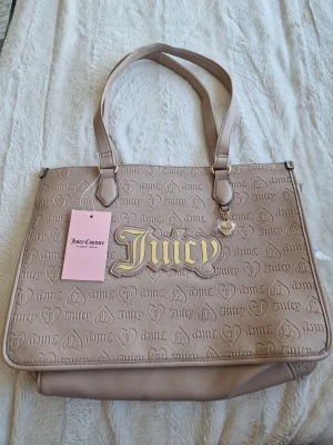 Beige shoppingväska Juicy Couture - Stilren shoppingväska i beige från Juicy Couture med präglat mönster och stort Juicy-tryck i guld på framsidan. Väskan har dubbla axelband, hjärtberlock och är tillverkad i konstläder. Perfekt för dig som vill ha en trendig och rymlig väska.