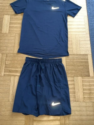 Svart  träningsset från Nike - Snyggt träningsset från Nike med en svart t-shirt och matchande shorts. T-shirten har en vit Nike-logga på bröstet och reflekterande detalj bak. Shortsen har resår i midjan, dragsko och vit logga på benet. Perfekt för gymmet eller en varm sommar dag 
