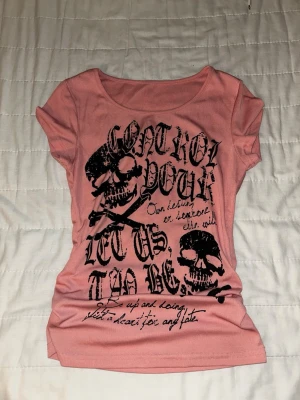 Rosa t-shirt med döskalletryck - Cool rosa t-shirt med svart döskalletryck och text framtill. T-shirten har rund halsringning och korta ärmar. Passformen är figurnära och materialet känns mjukt och stretchigt, perfekt för en edgy streetstil. Originalpris 115kr