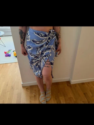 Blå sarong med vita blommor - Snygg strand-sarong i blått med stora vita blommor och knytning i sidan. Perfekt att slänga på sig över bikinin på stranden. Lätt och luftig modell som ger en avslappnad sommarkänsla.