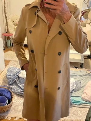 Klassisk beige trenchcoat - Stilren beige trenchcoat med dubbelknäppning och svarta knappar. Jackan har två fickor framtill och en klassisk krage. . Materialet känns slätt och följsamt.storlek är 34 ,jag som har medium i storlek passar även mig , som ni ser på bild .så jag kan  säga storlek är som mellan smal och medium 