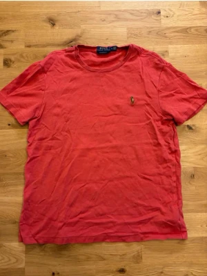 Röd t-shirt från Polo Ralph Lauren - Röd t-shirt från Polo Ralph Lauren med klassisk rund hals och broderad logga på bröstet. Modellen är custom slim fit och tillverkad i mjuk bomull. Perfekt för en avslappnad och stilren look.