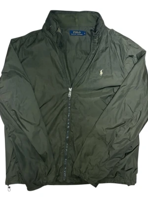Ralph Lauren jacka  - Snygg olivgrön vindjacka från Polo Ralph Lauren med dragkedja framtill och klassisk ståkrage. Jackan har det ikoniska broderade logotypen på bröstet och är tillverkad i lätt, vindtätt material. Perfekt för blåsiga dagar och enkel att matcha med olika outfits.