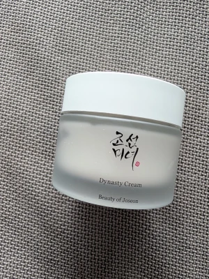 Dynasty Cream från Beauty of Joseon - En elegant ansiktskräm i en frostad, rund burk med vit lock. Dynasty Cream innehåller 2% niacinamid och 2% squalane för att återfukta och balansera huden. Minimalistisk design med svart text och koreanska tecken på framsidan.