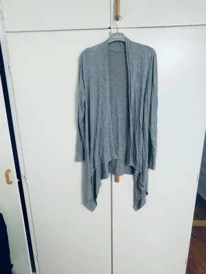 Grå lång kofta  - Säljer en tunn och mjuk grå kofta . Koftan har ett öppet och avslappnat fall med längre framstycken och lång ärm. Perfekt att slänga på sig över en t-shirt eller linne för en chill look.