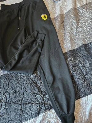 Svarta mjukisbyxor "Ferrari" - Säljer ett par svarta mjukisbyxor "Ferrari" med klassisk gul logga på vänster ben. Byxorna har ribbade muddar vid bensluten, dragsko i midjan och är gjorda i mjukt bomullsmaterial. Perfekta för chill dagar eller när du vill ha en sportig look.