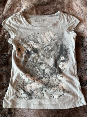 Grå t-shirt med tryck från MISS C - Snygg grå t-shirt från MISS C med coolt tryck av ett ansikte, stjärnor och blommor på framsidan. T-shirten har rund halsringning och korta ärmar. 