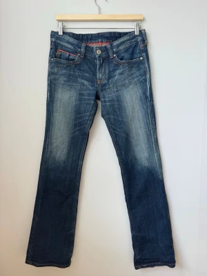 Blå bootcut jeans - Snygga blå bootcut jeans med slitningar och kontrastsömmar. Jeansen har fem fickor, röda detaljer och dekorativa bakfickor med knappar och röd söm. Klassisk denim med en cool tvätt och låg midja.