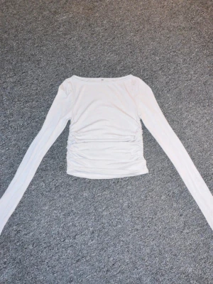 Gråvit långärmad topp med rynkade sidor - Säljer en gråvit långärmad topp med rynkade detaljer på sidorna. Toppen är figurnära i modellen. Perfekt att styla med jeans eller kjol för en clean look.