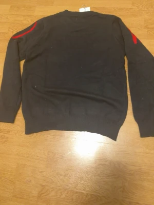 Svart stickad tröja med röd text - Svart stickad tröja från Valenti med stora röda bokstäver över bröstet och axlarna. Tröjan har rund halsringning, långa ärmar och ribbstickade muddar vid ärmslut och nederkant. Perfekt för dig som gillar en stilren men ändå iögonfallande look.