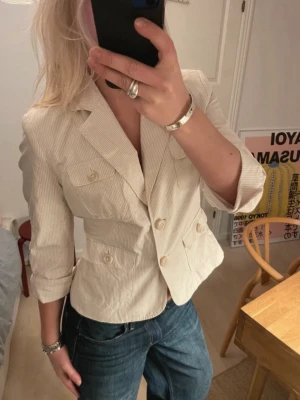 Beige kavaj  - Snygg beige kavaj från Merona med diskreta ränder, fyra fickor framtill och trekvartsärmar. Figursydd modell med klassisk krage och knäppning framtill. Perfekt att styla med jeans för en cool look.