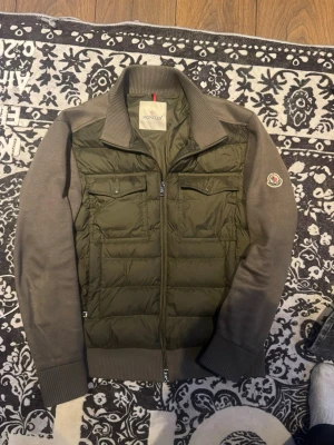 Olivgrön cardiganjacka från Moncler - Snygg olivgrön cardiganjacka från Moncler med quiltad front och stickade ärmar. Jackan har två bröstfickor med lock, ribbade muddar och dragkedja framtill. Moncler-logga på ärmen ger en exklusiv touch. Perfekt för lager på lager och streetwear.