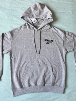 Grå hoodie från Gallery Dept. - Snygg ljusgrå hoodie från Gallery Dept. med svart tryck på bröstet. Klassisk modell med huva, dragsnören och stor magficka. Tillverkad i mjukt bomullsmaterial som är skönt mot huden. Perfekt för en avslappnad streetwear-look.