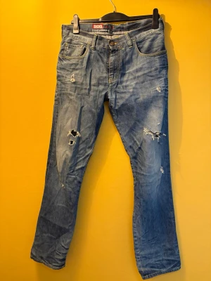 Blå slitna jeans från SICKO - Säljer ett par blå jeans från SICKO med flera slitningar och hål för en riktigt edgy look. 