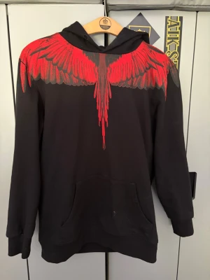 Svart hoodie med röd vingdesign - Säljer en svart hoodie från Marcelo Burlon med ikoniskt rött vingtryck över axlar och bröst, som fortsätter på ryggen. Hoodien har huva och känguruficka framtill. Tillverkad i mjuk bomull för maximal komfort och streetwear-vibe. M passar L