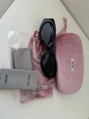 Svarta solglasögon från Miu Miu - Snygga svarta solglasögon från Miu Miu med markerad, kantig båge och guldiga detaljer vid gångjärnen. Kommer med rosa fodral i sammet, tygpåse och putsduk. Perfekt accessoar för att lyfta din stil.