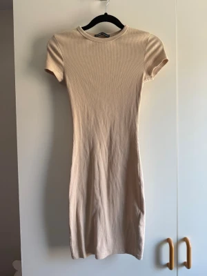 Beige ribbad kort klänning - En enkel och stilren beige kort klänning med ribbad struktur och rund hals. Klänningen har korta ärmar och en figurnära passform som ger en snygg siluett. Perfekt att styla med sneakers eller boots för en trendig look. Obs katt finns i hem!