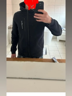 Svart hoodie från Sail Racing - Svart hoodie från Sail Racing med dragkedja hela vägen, huva med dragsko och en bröstficka med dragkedja. Snygg logga på bröstet och mjukt material som känns skönt mot huden. Perfekt för en avslappnad och sportig stil.