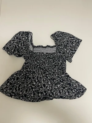 Svart blommig blus med puffärm - Supersöt svart blus med vitt blommönster och smockad midja. Toppen har korta puffärmar och en fyrkantig ringning som ger en romantisk vibe. Perfekt att styla med jeans eller kjol för en trendig look.