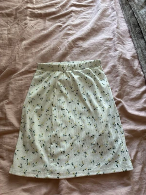 Vit blommig kortkjol -  vit kortkjol med små blommor i blått och gult. Kjolen har resår i midjan för en bekväm passform och är tillverkad i ett mjukt, ribbat material. Använt få gånger 
