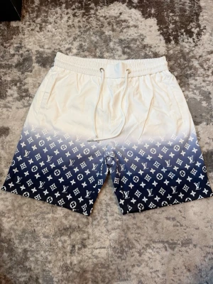 Louis Vuitton vita och blå shorts - Snygga shorts från Louis Vuitton med vit ovandel som övergår i mörkblå nederdel med klassiskt LV-mönster. Shortsen har snörning i midjan och är perfekta för sommaren. Materialet känns lätt och bekvämt.
