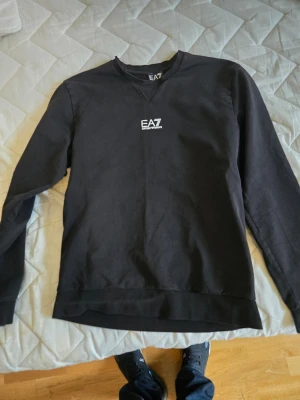 Svart sweatshirt från EA7 Emporio Armani - Svart sweatshirt med rund hals från EA7 Emporio Armani. Tröjan har en stilren design med vitt EA7-tryck på bröstet och ribbade muddar vid ärmslut och nederkant. Perfekt för en avslappnad och trendig look.