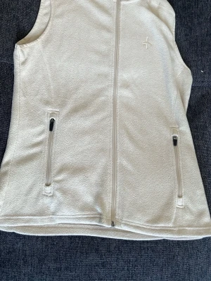 Beige fleeceväst från Cross Sportswear - Beige fleeceväst från Cross Sportswear med hel dragkedja framtill och två fickor med dragkedja. Västen har en stilren design och är perfekt för lager-på-lager. Broderad logga på bröstet.