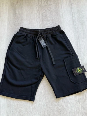 Svarta shorts från Stone Island - Svarta shorts från Stone Island med elastisk midja och snörning. Stora fickor på sidorna och klassisk logotyp-patch på ena benet. Perfekta för en avslappnad stil.