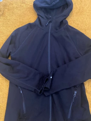 Svart hoodie med dragkedja - Svart hoodie med hel dragkedja framtill och två fickor med dragkedja. Klassisk modell med huva och långa ärmar. Perfekt för en avslappnad stil och enkel att matcha med andra plagg.