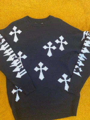 Svart stickad tröja med vita kors - Cool svart stickad tröja med vita korsmönster över hela tröjan och ärmarna. Tröjan har rund halsringning och ribbstickade muddar vid ärmslut och nederkant. Perfekt för dig som gillar streetwear och unika detaljer.