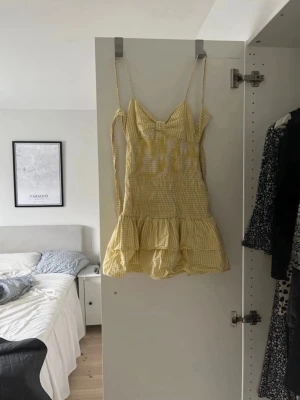 Gul rutig kort klänning med volang - Söt gul och vit rutig kort klänning med smala axelband, volangkant nertill och knytning i ryggen. Perfekt för varma dagar och har en lätt, luftig känsla. Klänningen har en feminin passform och är tillverkad i ett mjukt bomullstyg. Kan oxå byta klänningen mot en likadan fast i storlek S🥹🥹