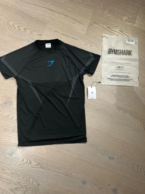 Svart träningströja från Gymshark - Snygg svart träningströja från Gymshark med gråa grafiska detaljer och blå logga på bröstet. Tillverkad i ett lätt och snabbtorkande funktionsmaterial, perfekt för intensiva träningspass. Kortärmad och med modern passform.