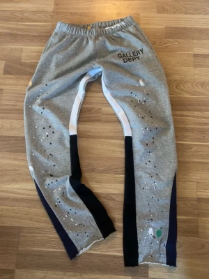 Grå mjukisbyxor från Gallery Dept. - Snygga grå mjukisbyxor från Gallery Dept. med svart logga på vänster höft. Byxorna har elastisk midja, vita och svarta detaljer längs benen samt färgstänk och hål för en unik, streetinspirerad look. Perfekta för dig som gillar trendiga och bekväma plagg. Priset går att diskuteras!