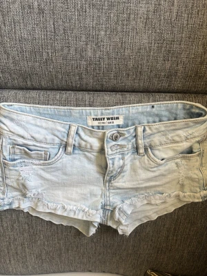 Ljusblå jeansshorts från Tally Weijl - Säljer ett par ljusblå jeansshorts från Tally Weijl med fransig kant och slitna detaljer. Shortsen har fem fickor, bälteshällor och knappgylf. Perfekta för varma dagar och ger en avslappnad vibe.