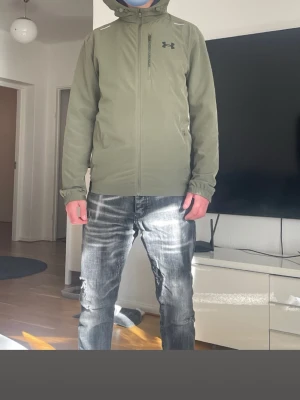 Grön vindjacka från Under Armour - Snygg grön vindjacka från Under Armour med dragkedja framtill och hög krage. Jackan har en bröstficka med dragkedja och diskret logga på bröstet. Tillverkad i lätt och tåligt material, perfekt för blåsiga dagar. Justerbar huva och muddar för extra komfort.