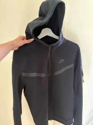 Svart Nike teech hoodie med dragkedja - Säljer en svart hoodie från Nike med hel dragkedja och huva. Tröjan har en diskret Nike-logga på bröstet och en snygg detalj med en horisontell rand över bröstet. Ärmarna har en ficka med dragkedja. Perfekt för en sportig och avslappnad stil.
