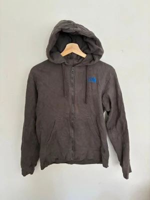 Grå hoodie från The North Face - Säljer en grå hoodie från The North Face med dragkedja och blå logga på bröstet. Hoodien har huva med dragsko och två stora fickor framtill. Perfekt för en avslappnad och cool stil. Storlek S