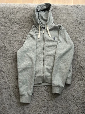 Grå hoodie från Polo Ralph Lauren - Snygg grå zip-hoodie från Polo Ralph Lauren med klassisk broderad logga på bröstet. Hoodien har dragsko i huvan, ribbade muddar och fickor framtill. Perfekt för en chill och stilren look.